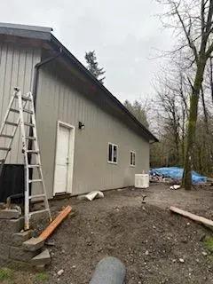 Gray building exterior with ladder, gravel yard, and blue tarp in a wooded area