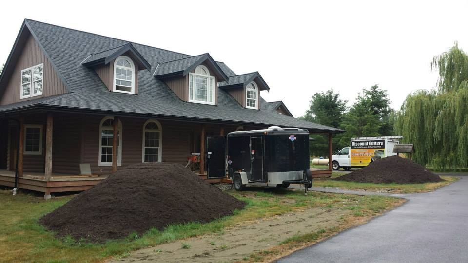 House with dark roof beside piles of dirt and a black trailer during landscaping work