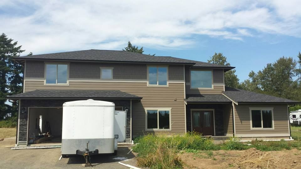 Two-story tan house under construction with a white trailer in the driveway and overgrown yard