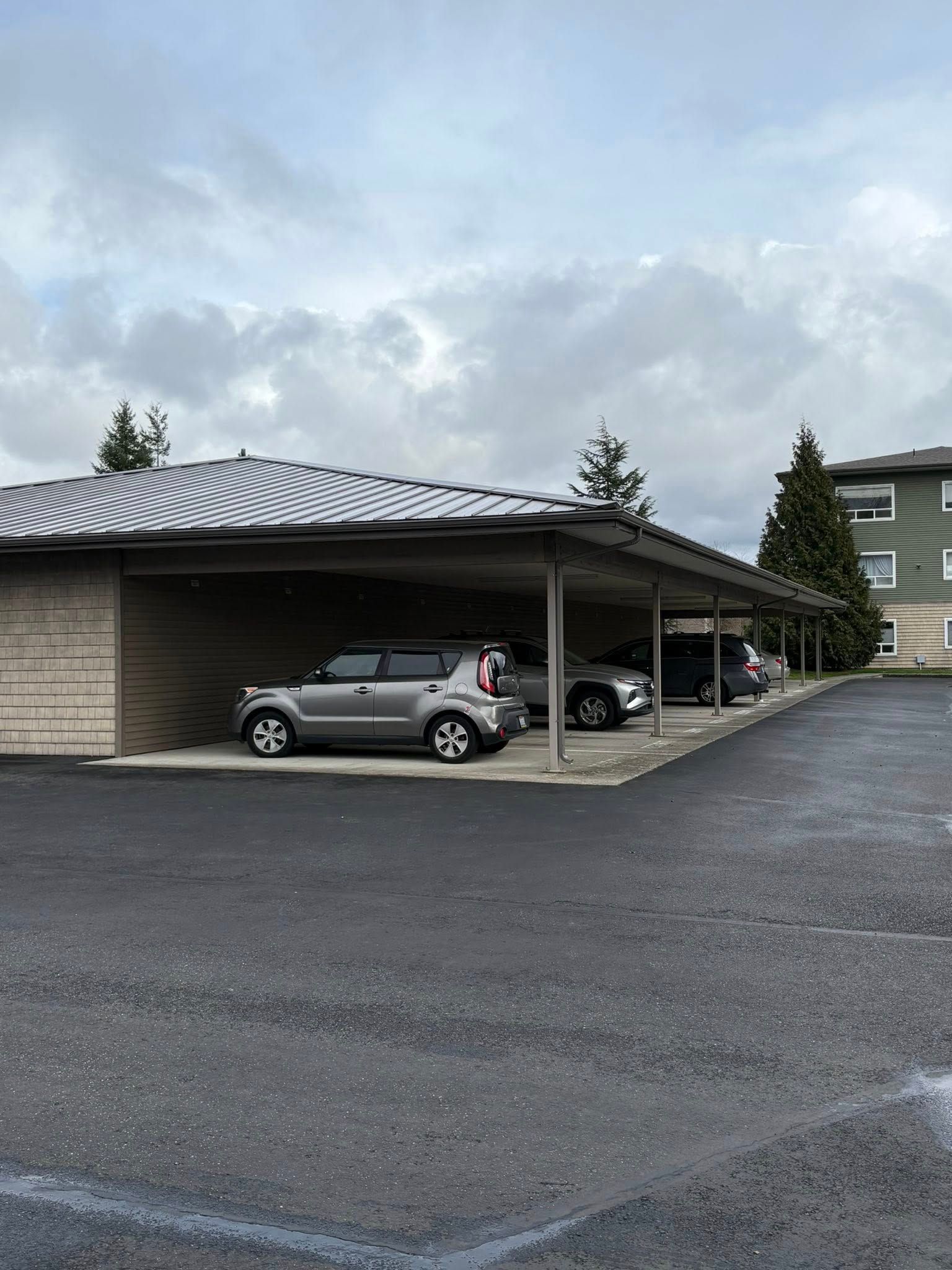 Covered parking area with a parked silver car beside a building under a cloudy sky.