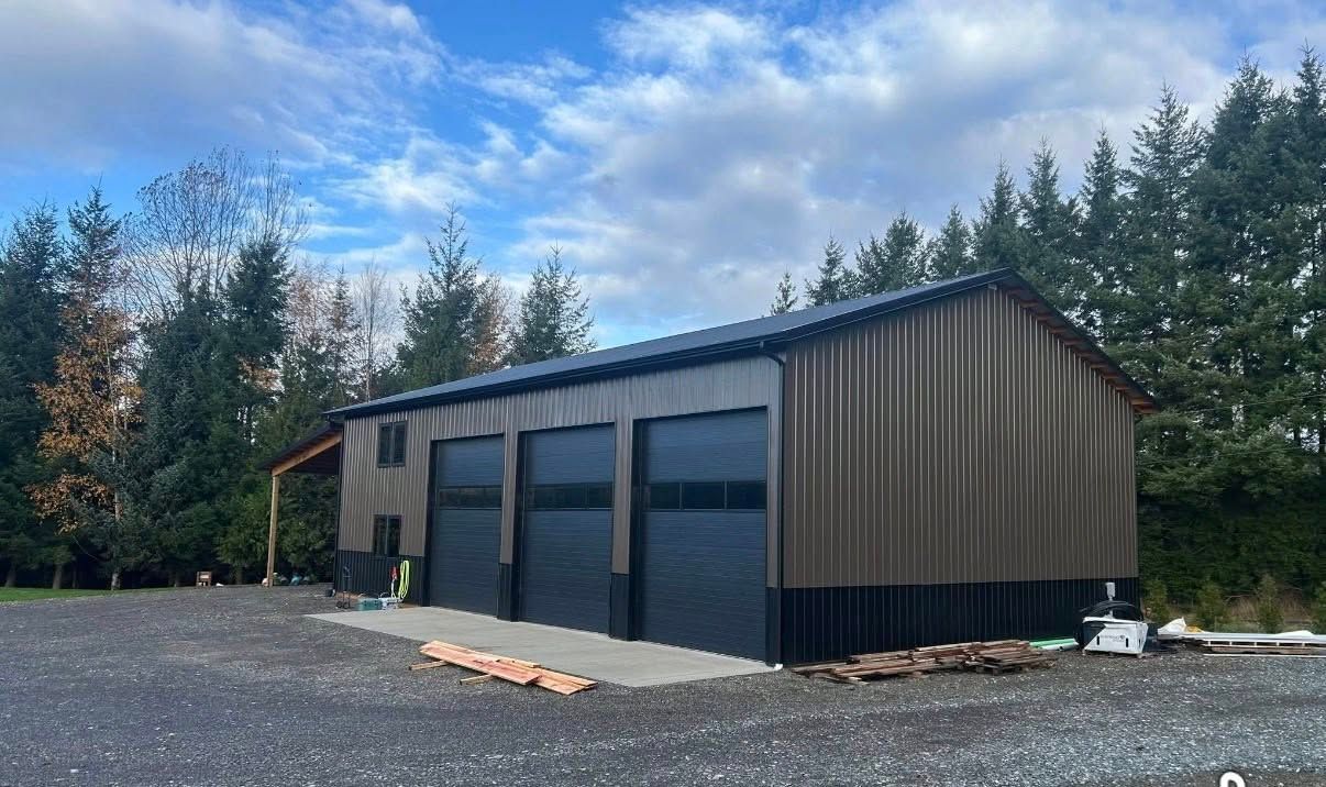 Modern metal workshop building with large garage doors on a gravel lot, surrounded by trees under a cloudy sky