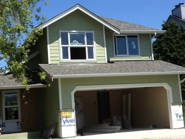 Two-story green house with open garage under renovation, white-trim windows, and blue sky.