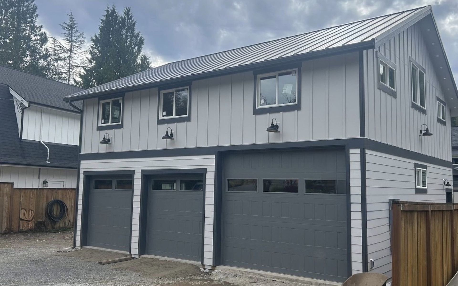 Gray two-story garage with three doors and upper windows beside a wooden fence under cloudy skies