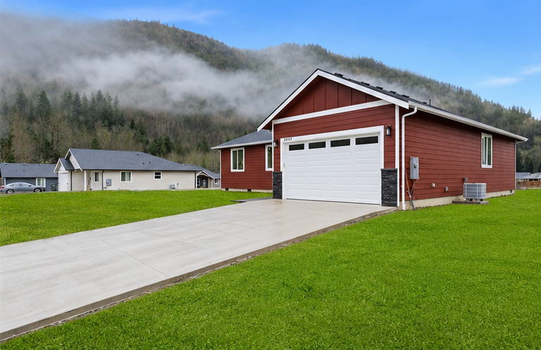 Red garage with white doors beside a concrete driveway and green lawn, backed by misty hills.