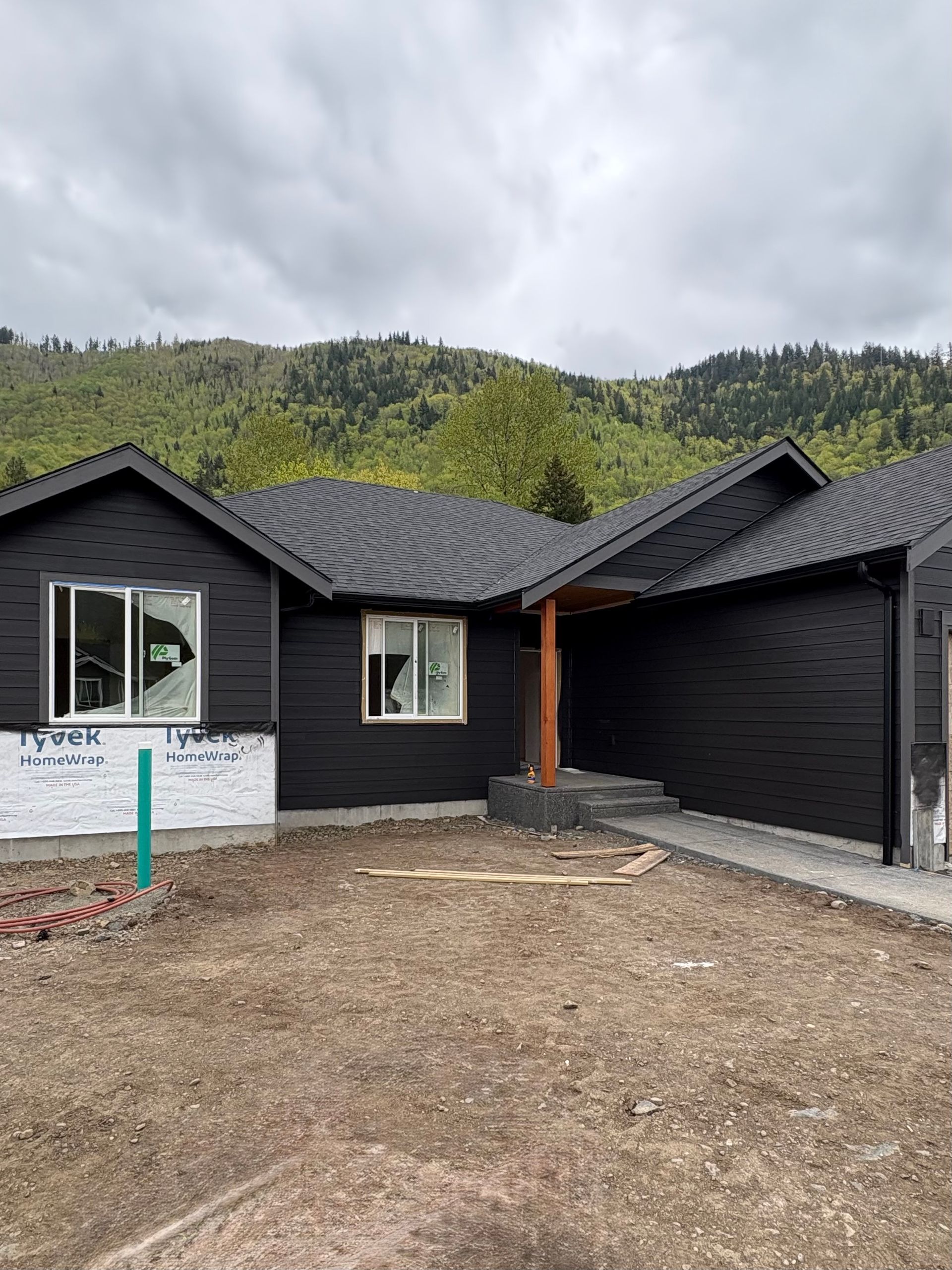 Black single-story house under construction with gravel yard and green hills in the background