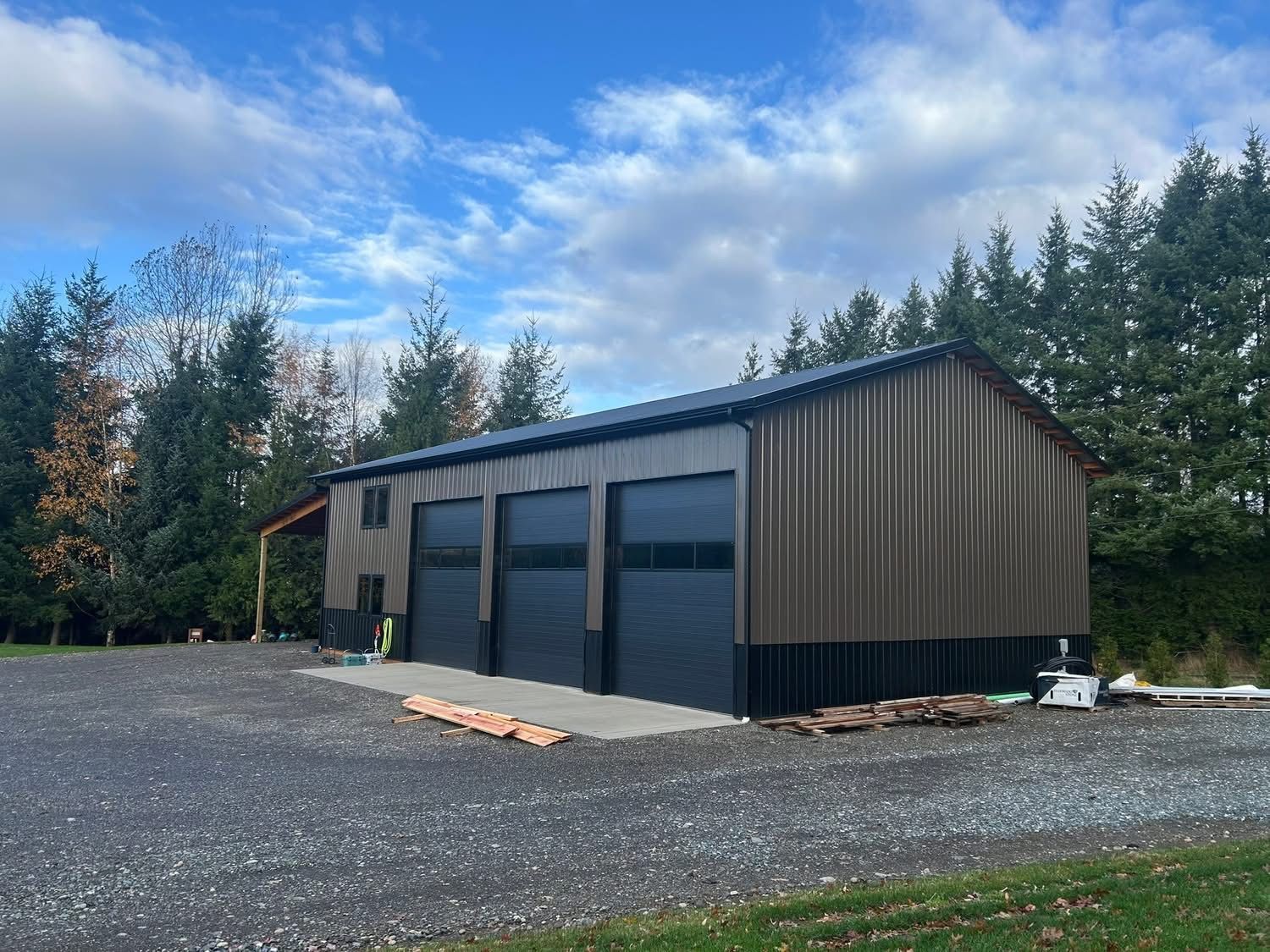 Small dark metal garage or workshop with blue doors on a gravel lot beside trees under a cloudy sky