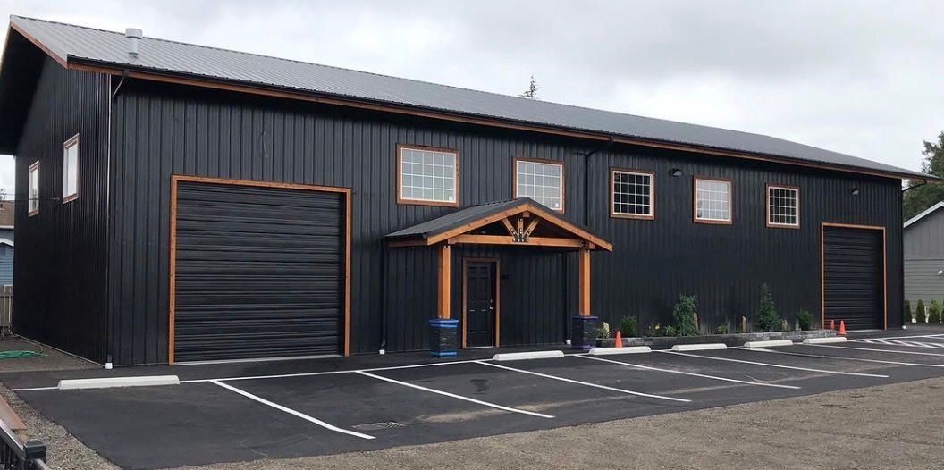 Black building with garage-style doors and a small wooden entrance canopy beside a parking lot