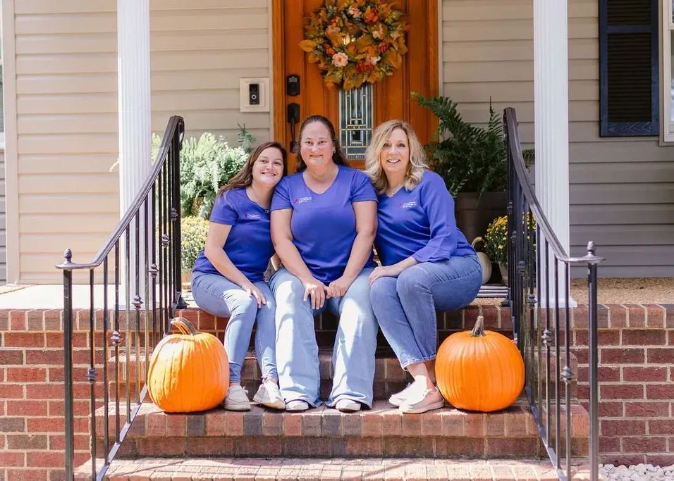 Three women in blue shirts and jeans sit on porch steps with pumpkins.