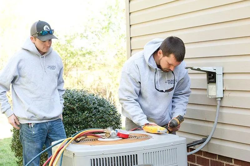 Two HVAC technicians working on an air conditioning unit outside a house. One is using a power tool.