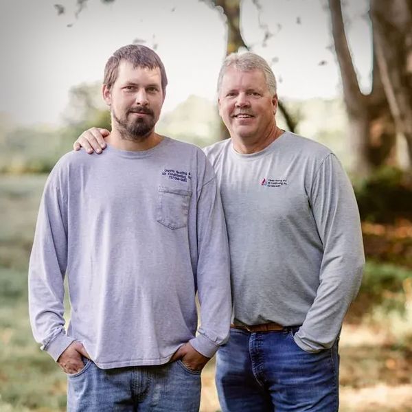 Two men in matching gray shirts and jeans, smiling, posing outdoors with an arm around each other.