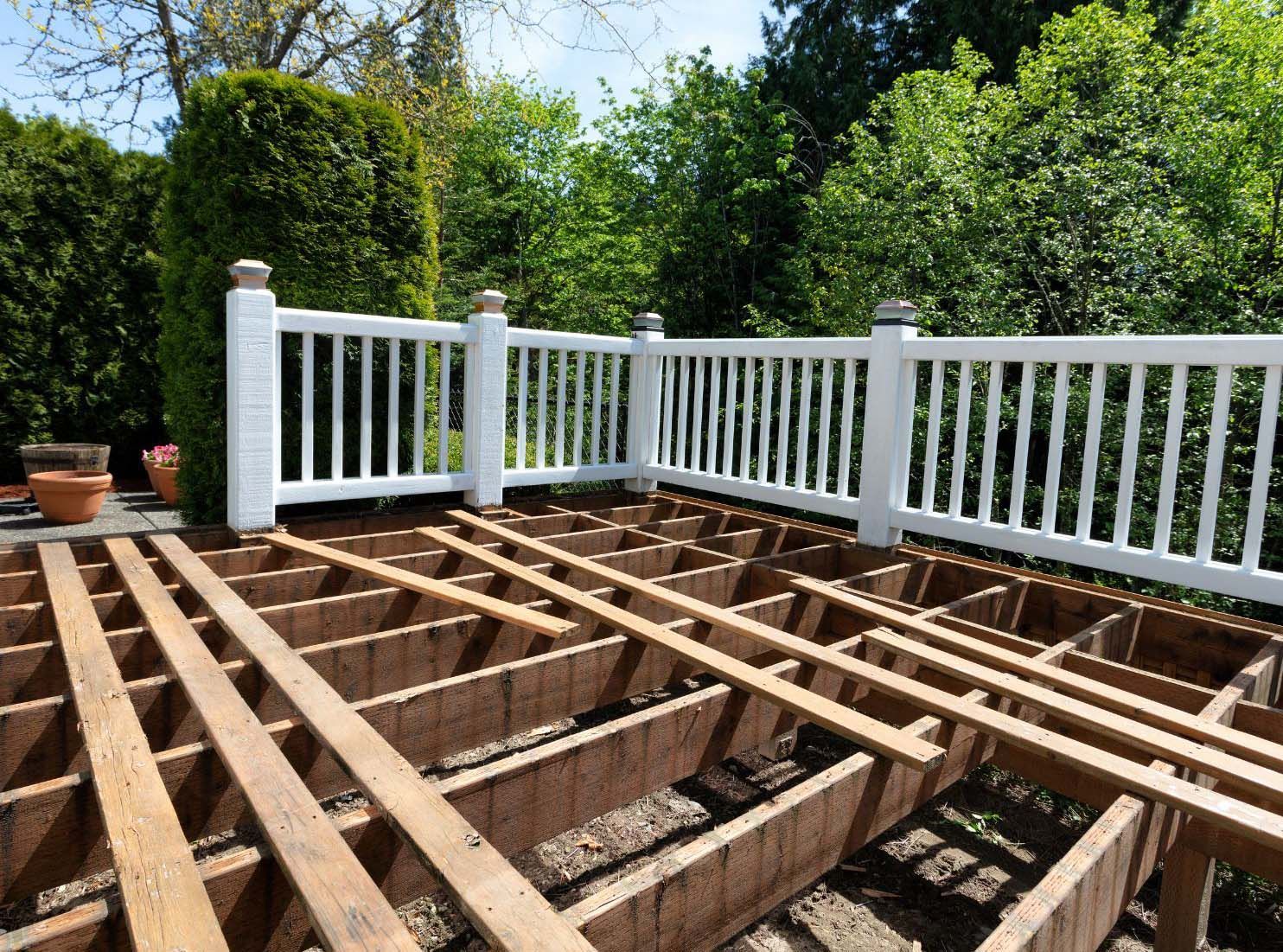 Deck under construction, exposed beams, white railing, green trees in background, sunny day.