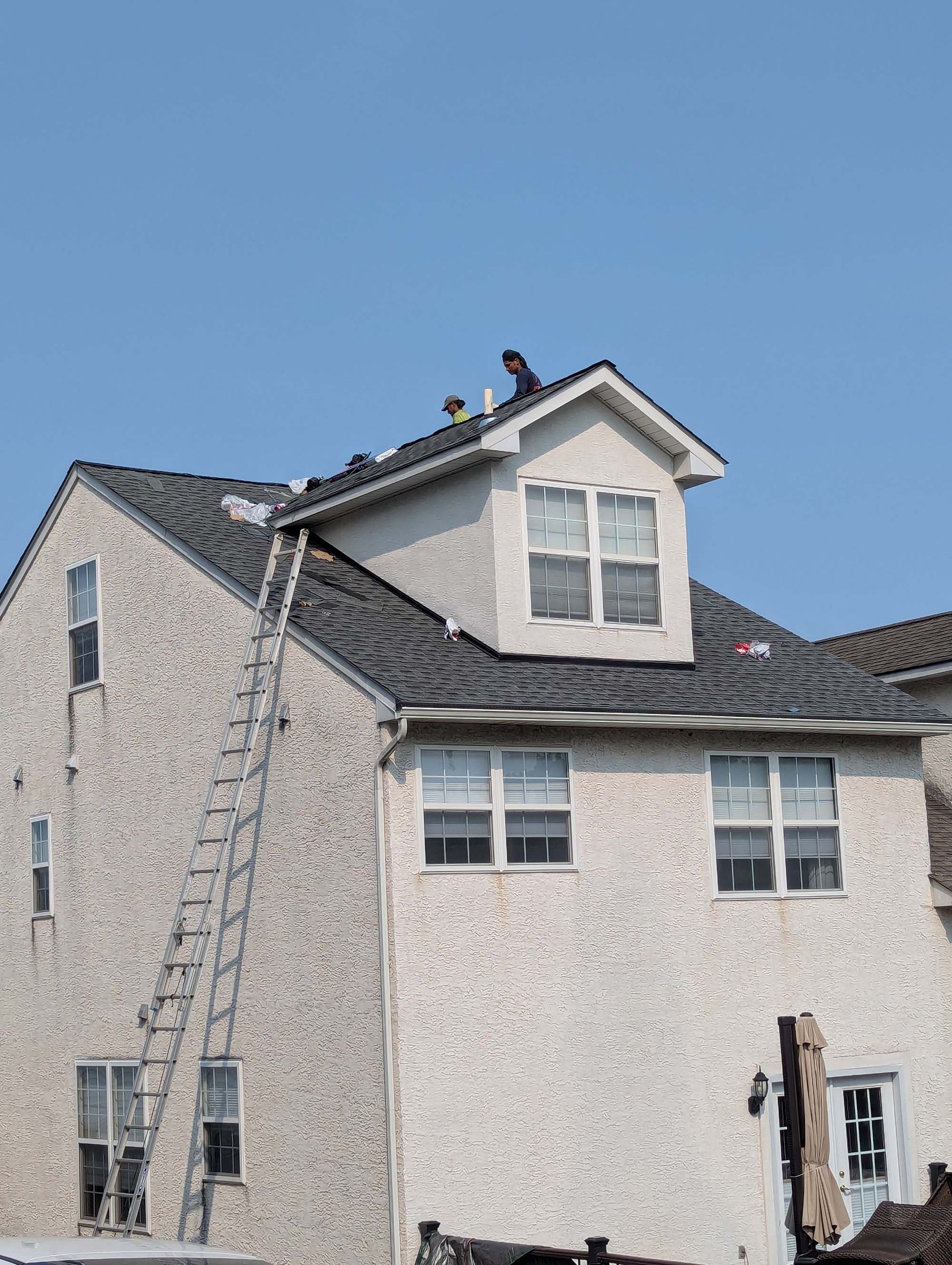 Workers on a roof with a ladder, blue sky, house with dormer and multiple windows.