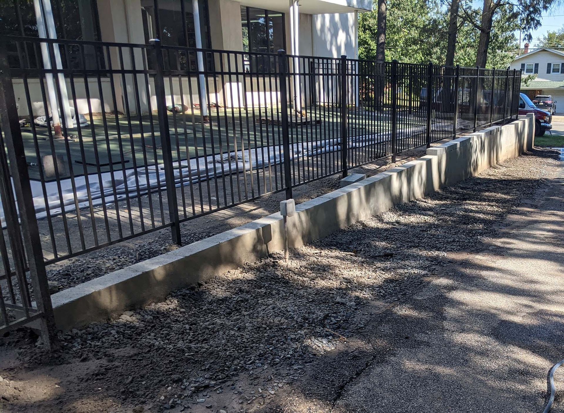 Black metal fence on a low concrete retaining wall, bordering a gravel-covered area, near a building.