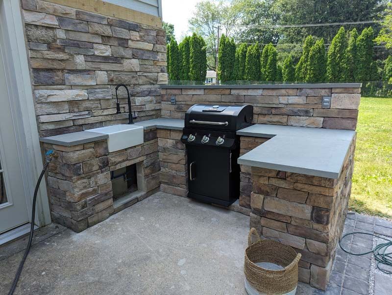 Outdoor kitchen with stone facing, concrete countertops, grill, sink, and neutral tones.