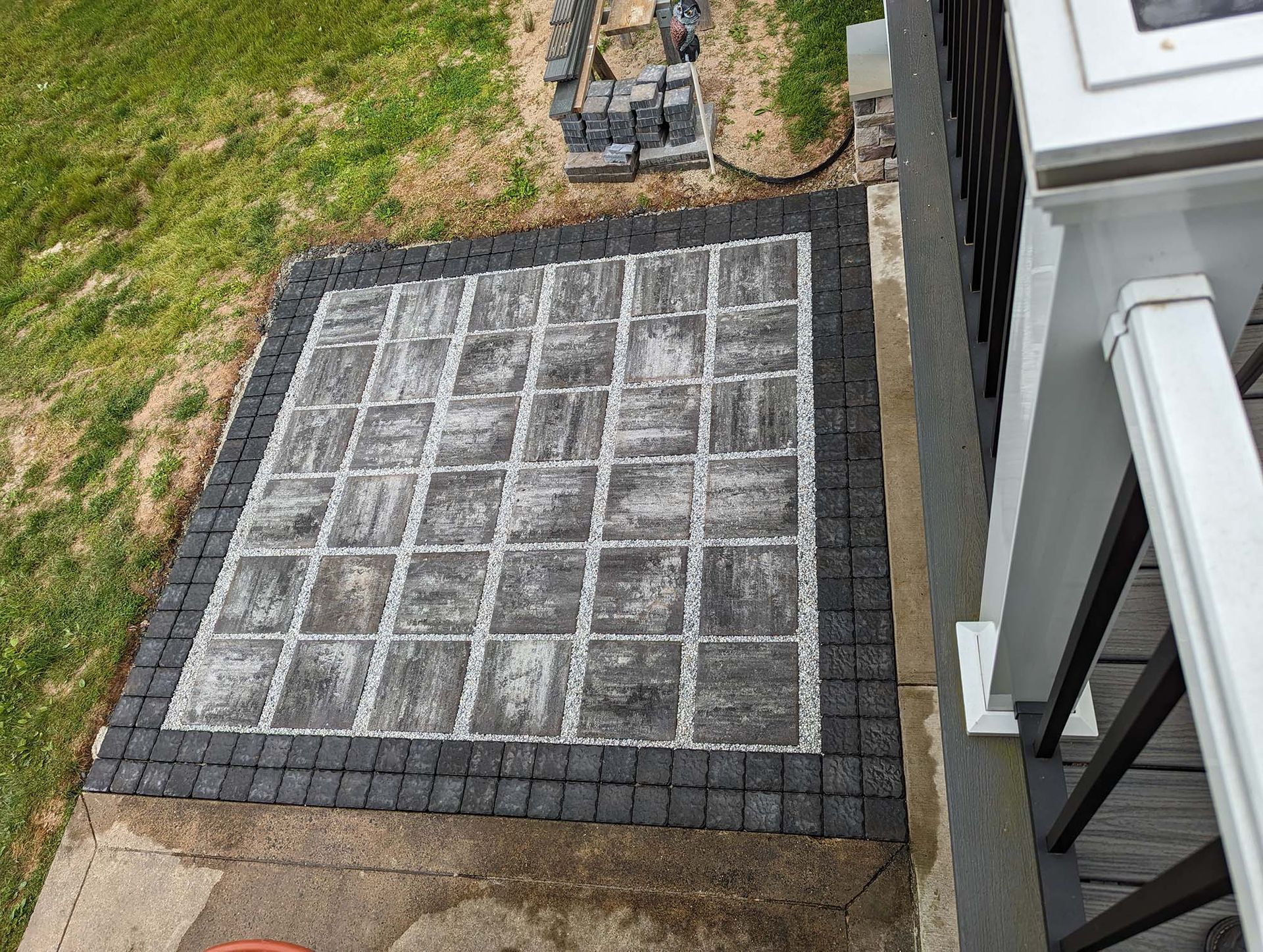 Paver patio with dark border next to a white porch post and railing; grass and pavers in background.