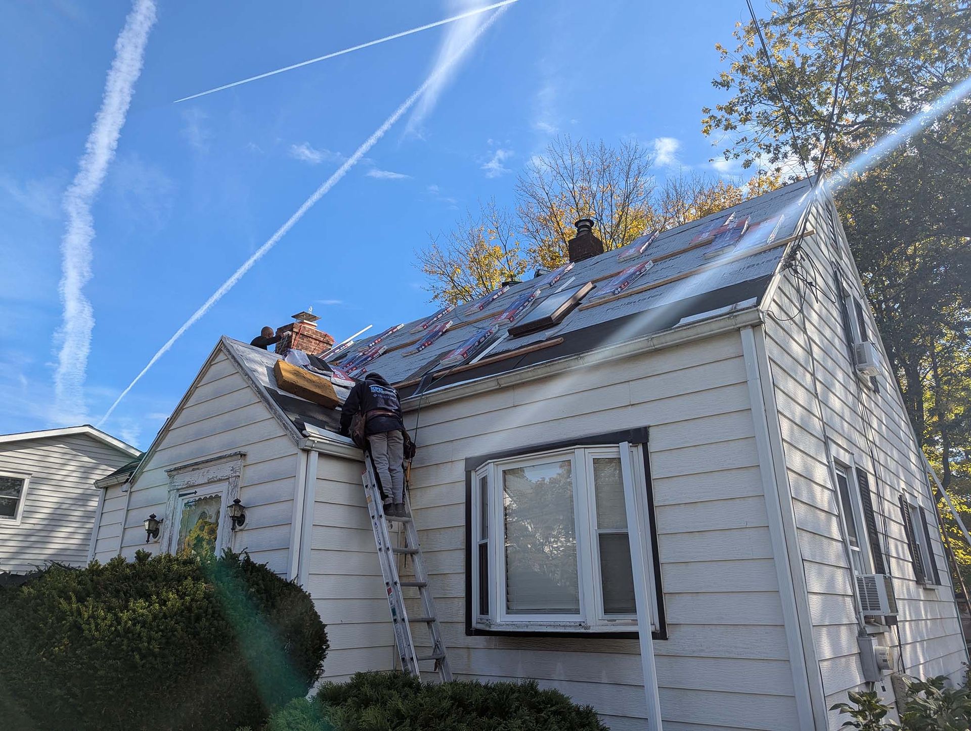 Workers on ladder installing roofing on a white house under a blue sky.