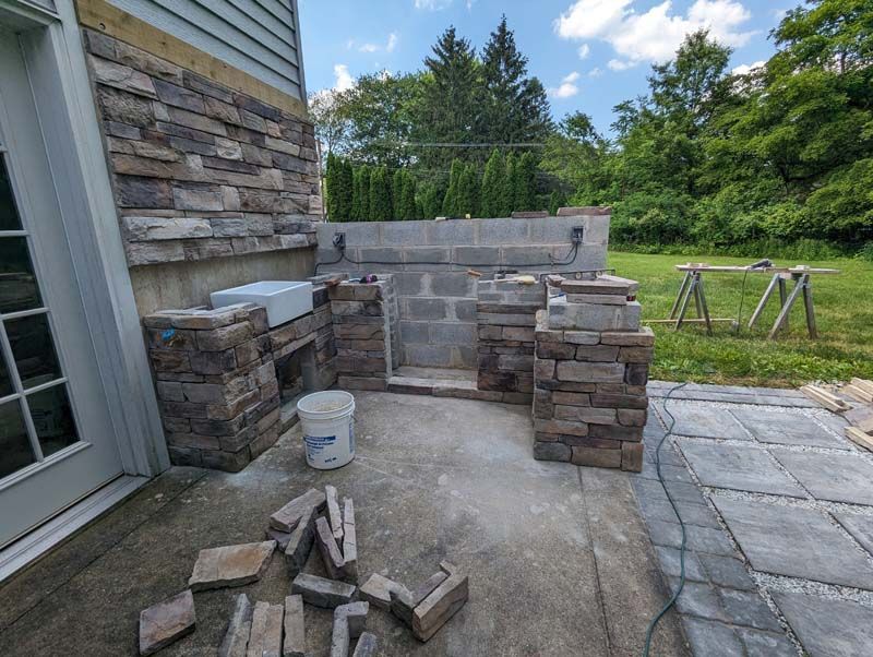 Outdoor stone kitchen under construction next to a house with paving stones and tools.
