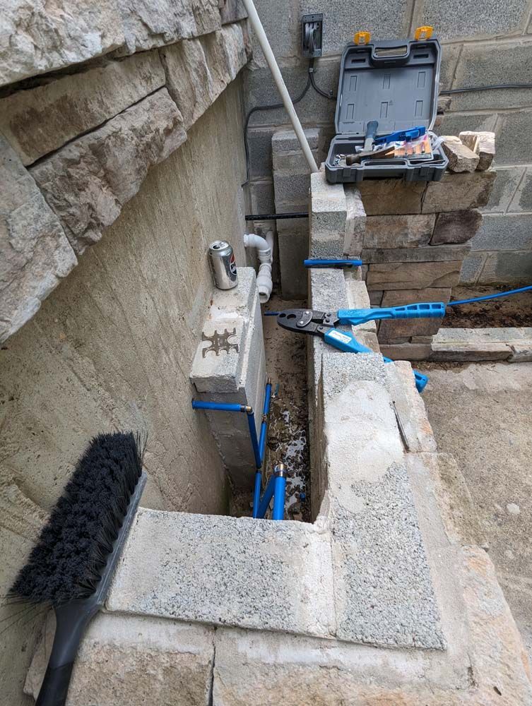 Construction site: blue water pipes in a stone wall trench. Tools, brush, and electrical box nearby.