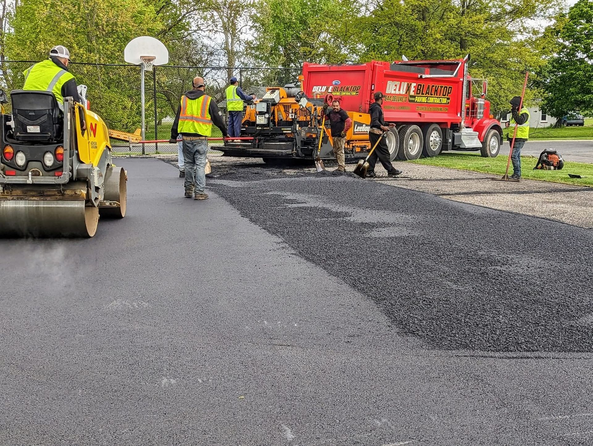 Asphalt paving crew working on a road, with truck, roller, and workers.