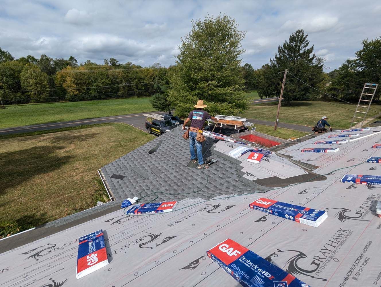 Roofers installing shingles on a house roof on a sunny day. Blue, red and white GAF packaging is visible.