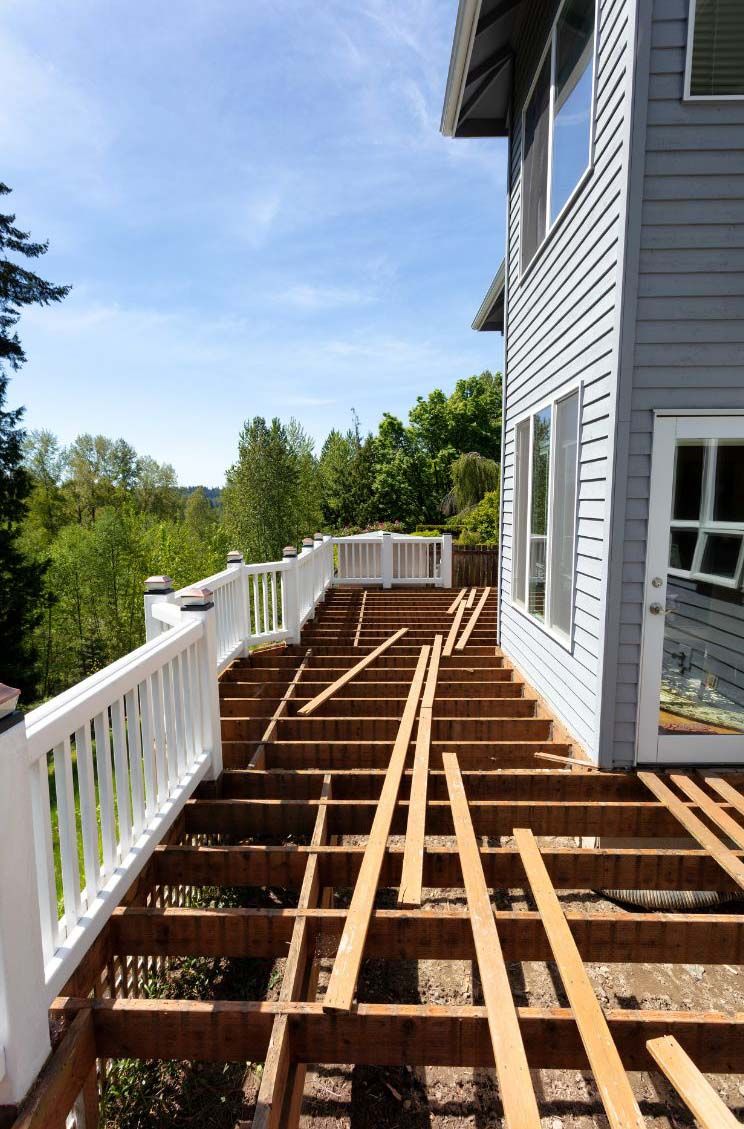 Deck under construction next to a two-story house with a white railing and trees in the background.