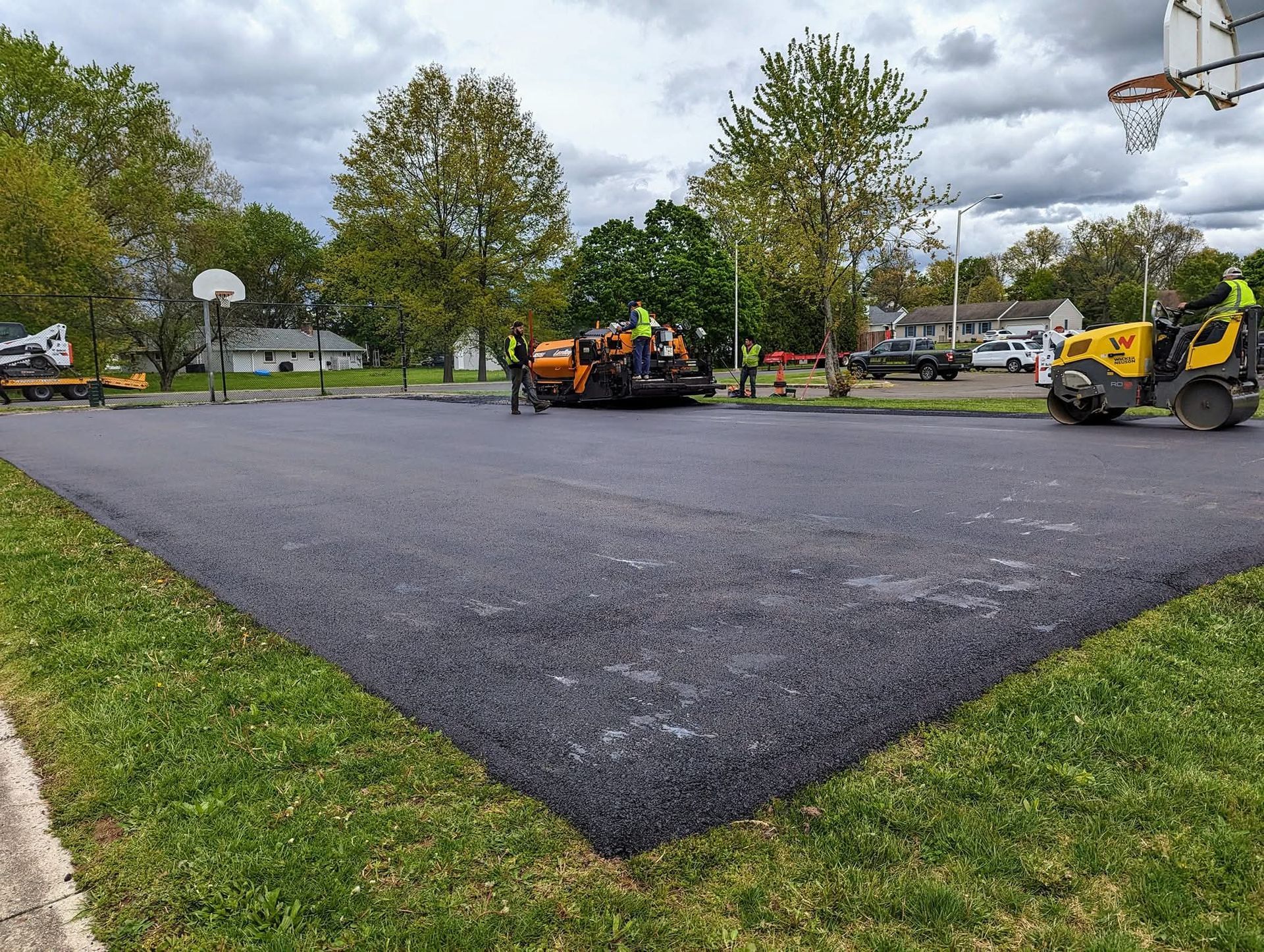 Asphalt paving crew resurfacing a basketball court; machinery, workers, and green grass.