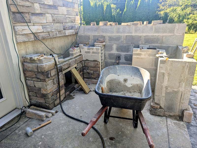 Construction of an outdoor brick structure. A wheelbarrow holds cement mix, with blocks and bricks on the patio.