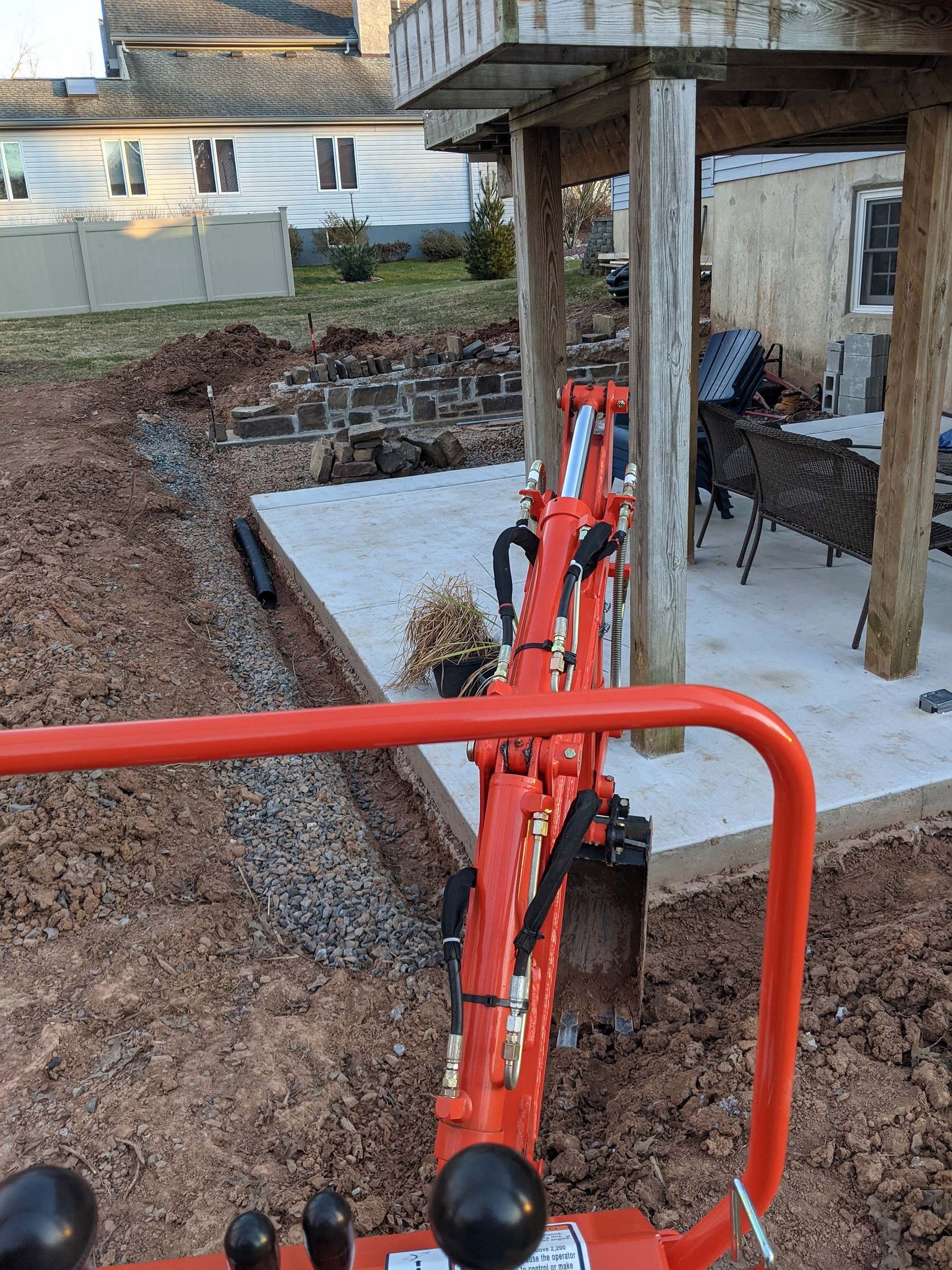 Orange excavator digging a trench beside a concrete patio, next to a wooden deck.