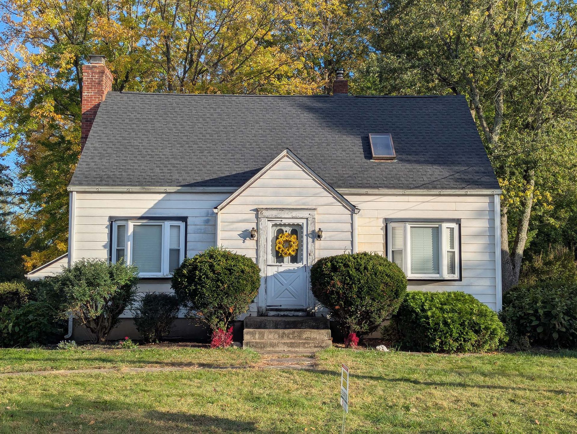 Small white house with bushes, windows, brick chimney, and wreath on the front door.