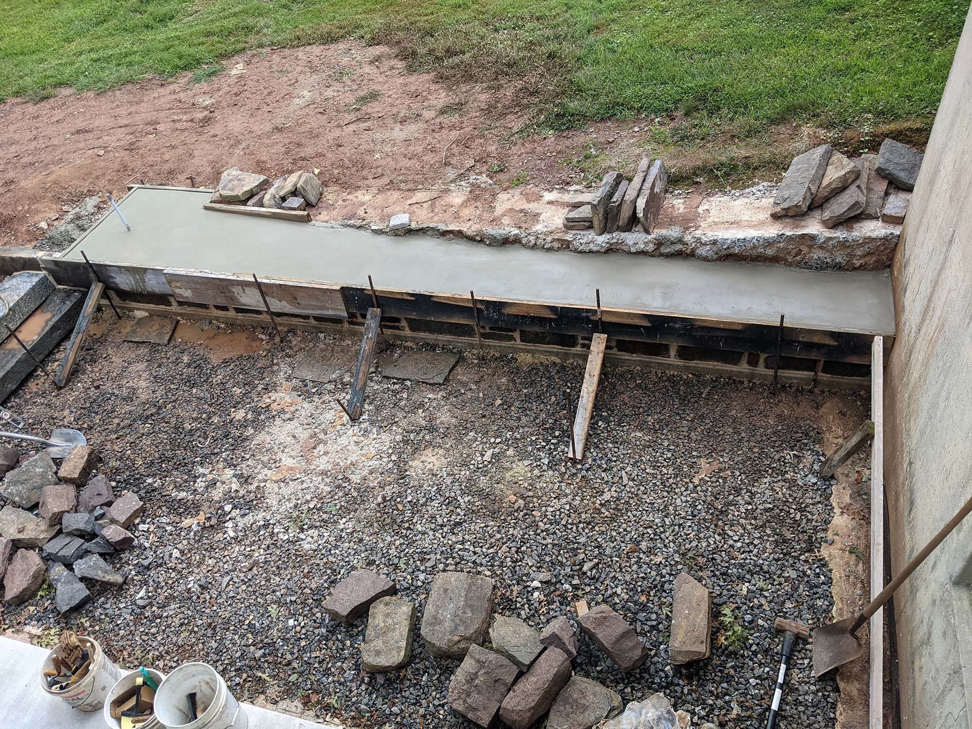 Construction site with fresh concrete border and gravel ground. Bricks and tools scattered around.