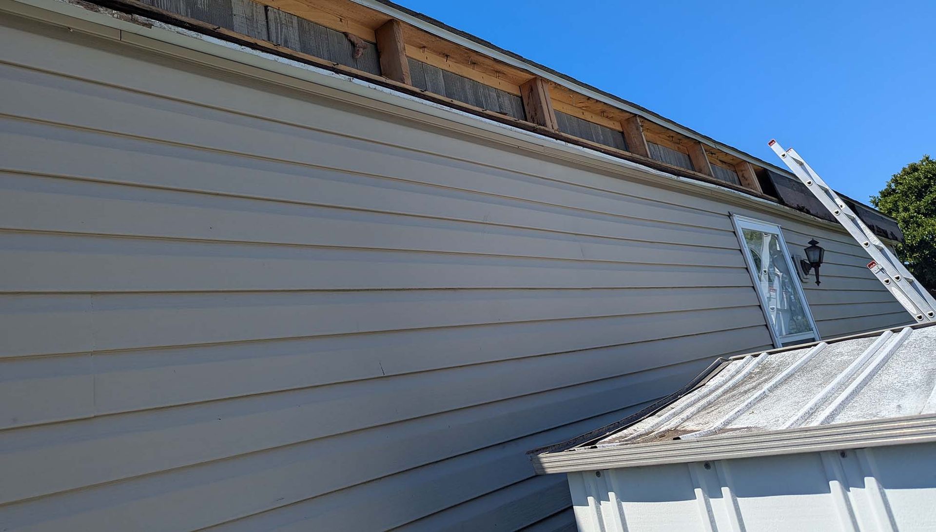 Side of a house with light siding and a ladder, against a clear blue sky.