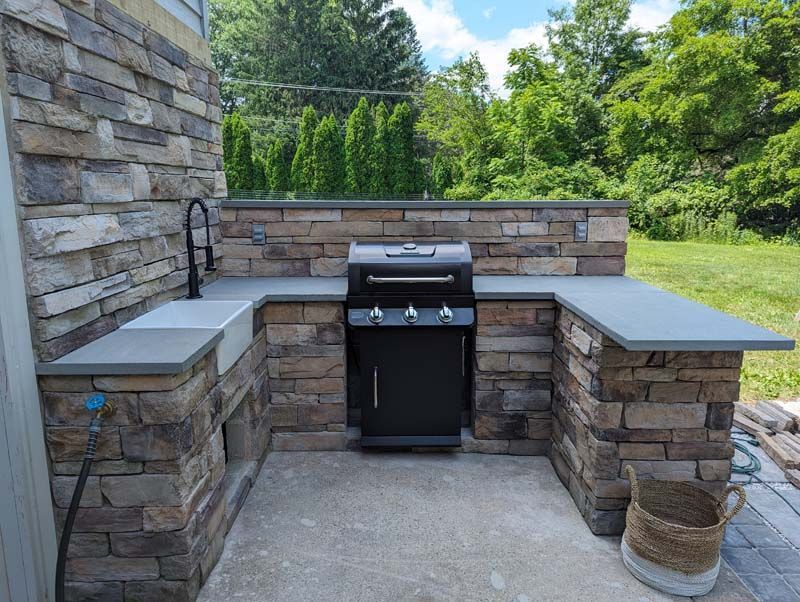 Outdoor kitchen with stone walls, gray countertop, black grill and faucet, and surrounding greenery.
