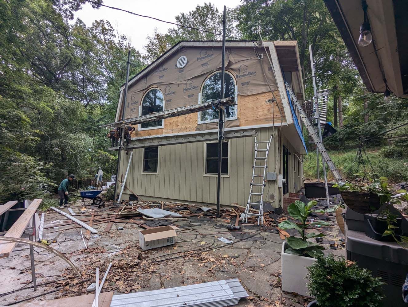 House under construction with scaffolding. Siding being replaced. Forest backdrop, scattered materials on ground.
