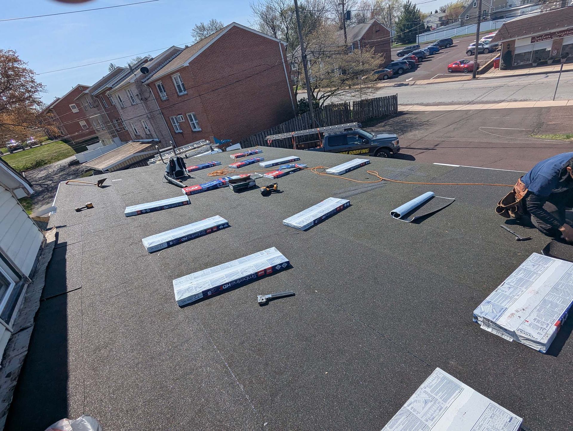 Workers installing something on a flat, black roof; tools, materials, and buildings in the background.