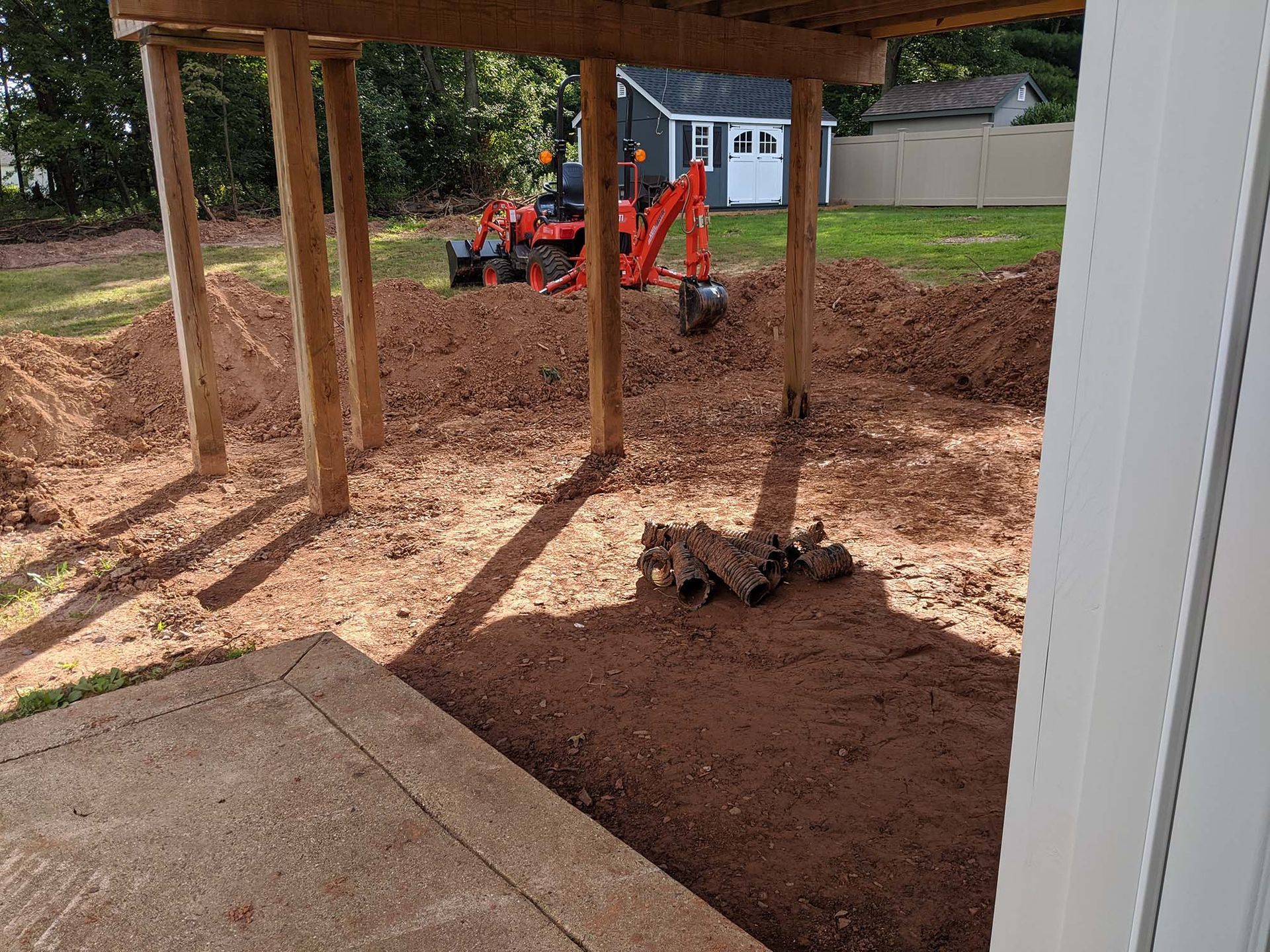 A small tractor digs dirt under a wooden deck. Brown soil, a house, and a shed are visible.