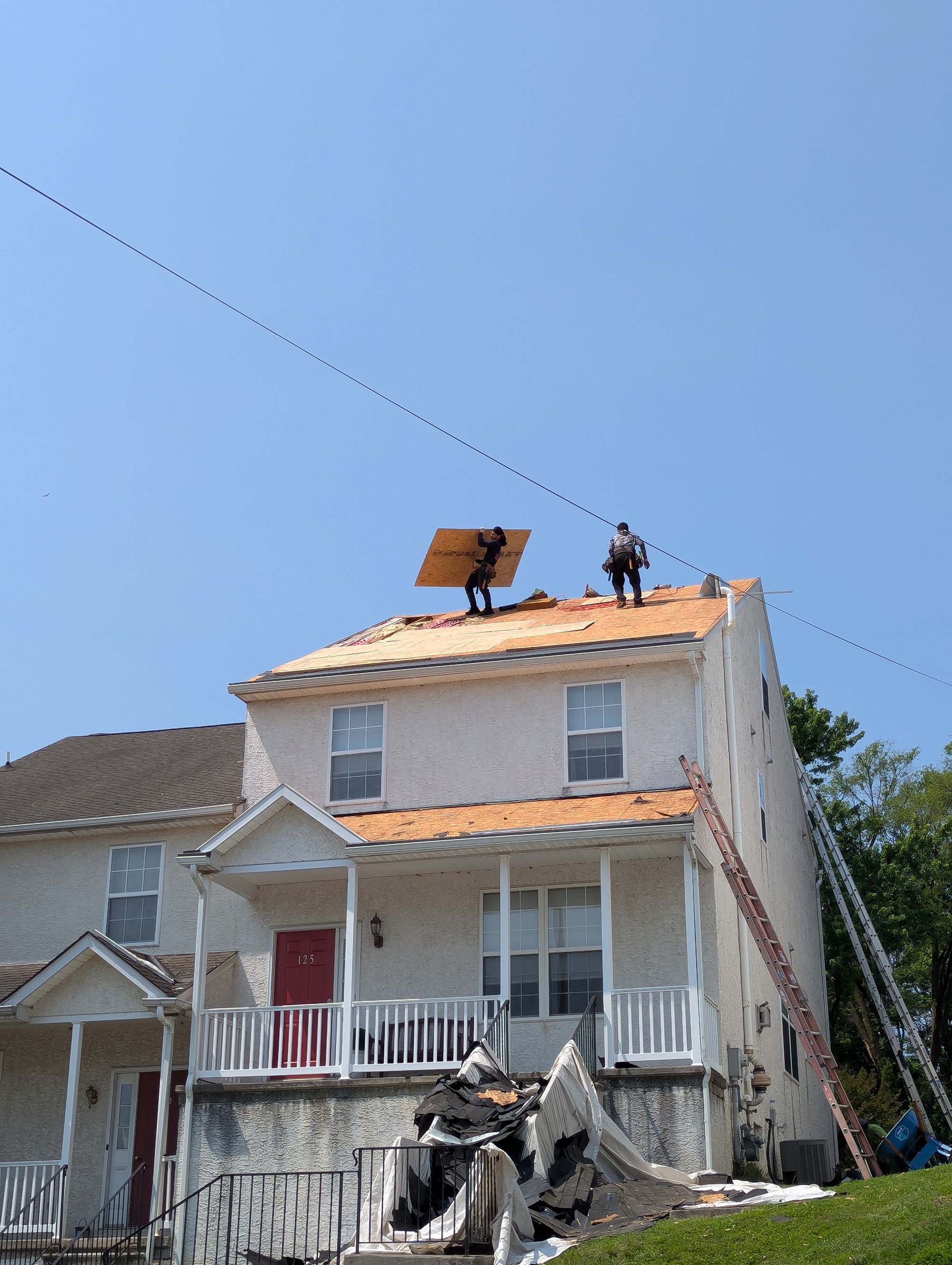 Two workers on a house roof. One holds a large square piece. Clear blue sky.