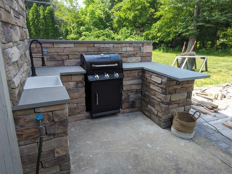 Outdoor kitchen with stone walls, gray countertops, grill, sink, and faucet.