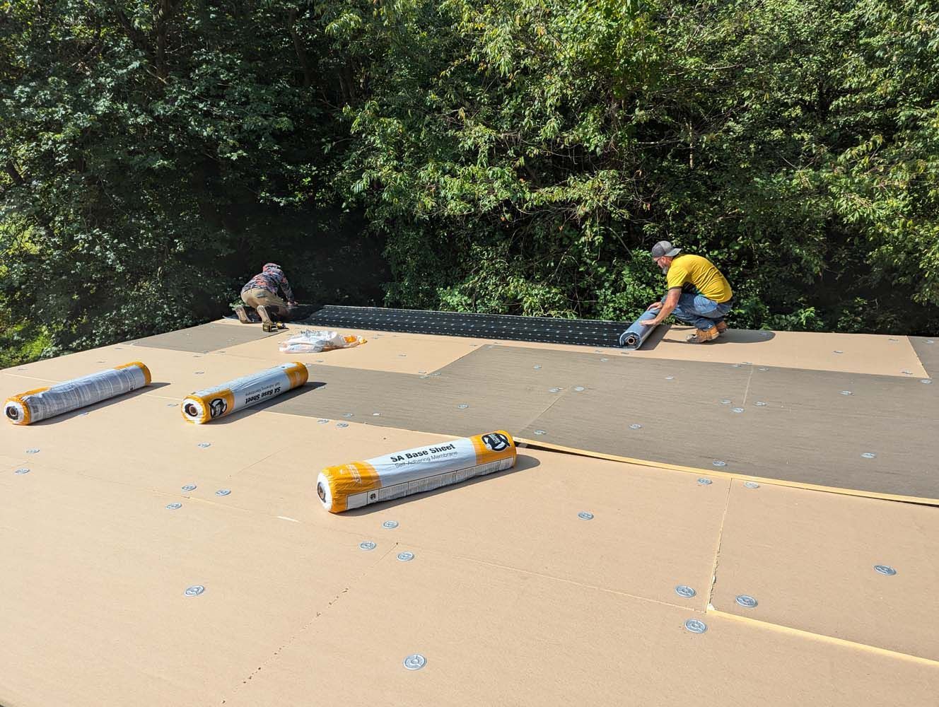 Two workers installing roofing materials on a roof with a forest backdrop; beige and black materials are in use.