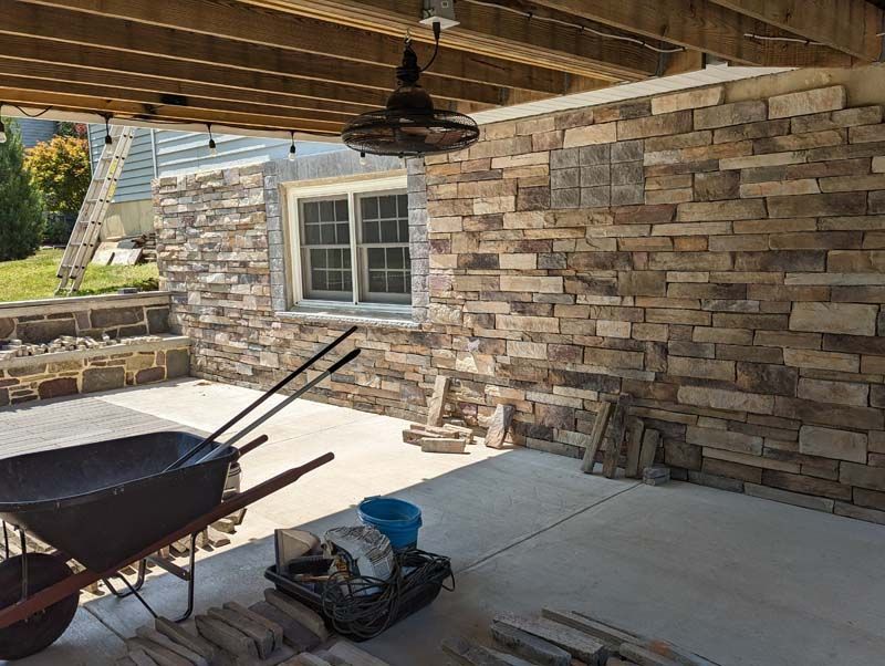 Stone facade installation on an outdoor patio with a wheelbarrow, tools, and a window.