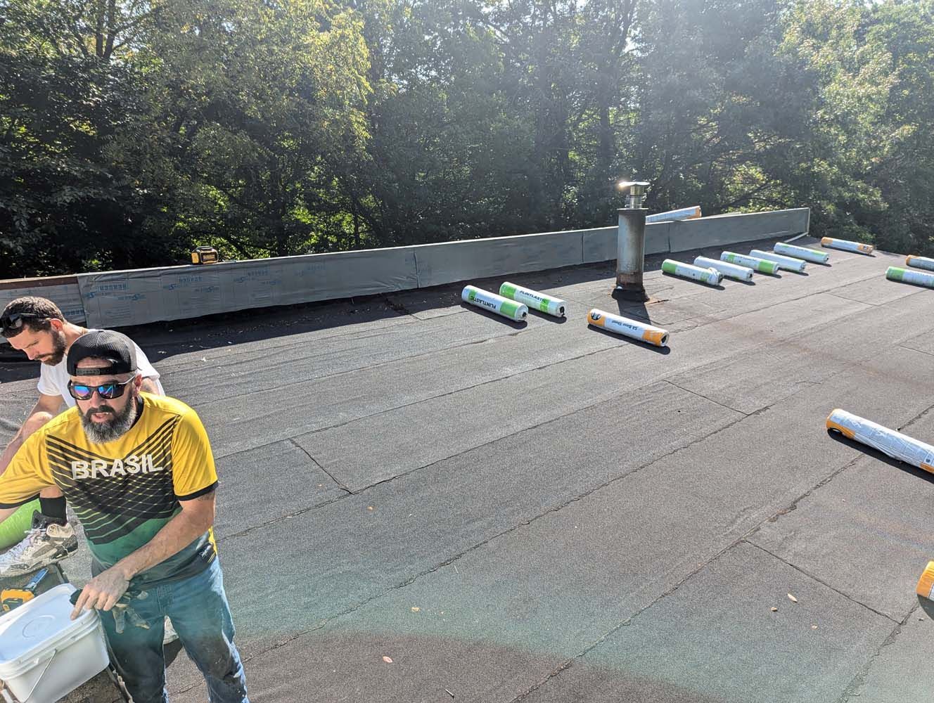 Two roofers working on a flat roof, surrounded by rolled roofing materials, sunny day.
