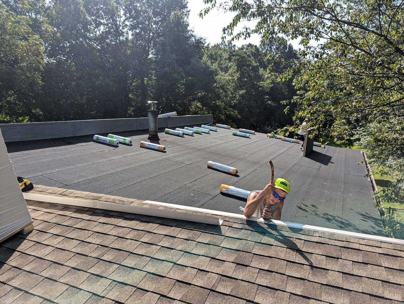 Person working on a flat roof with rolls of material, trees in background, sunny day.