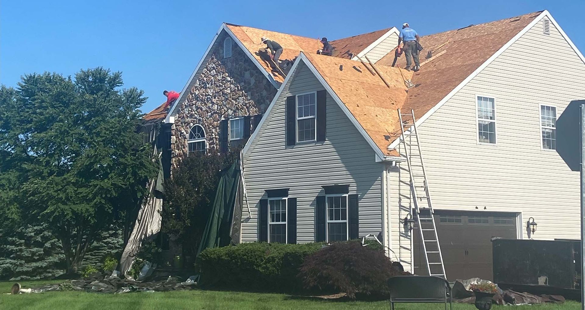 Roofers working on a two-story house with shingles missing, a ladder, and a partly shingled roof under a clear, blue sky.