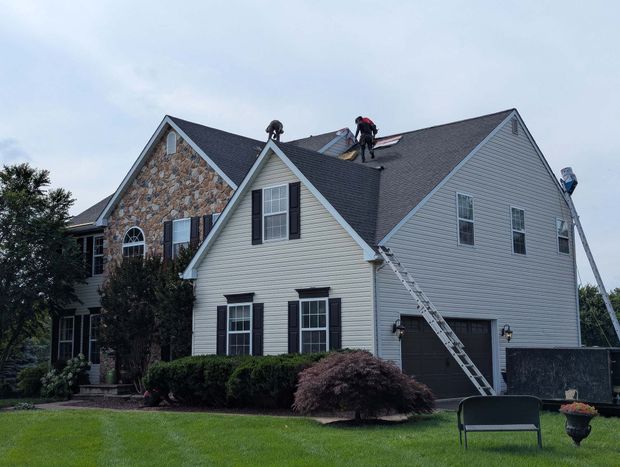 Two workers on roof of a two-story house with stone and siding. Ladders propped up, overcast sky.