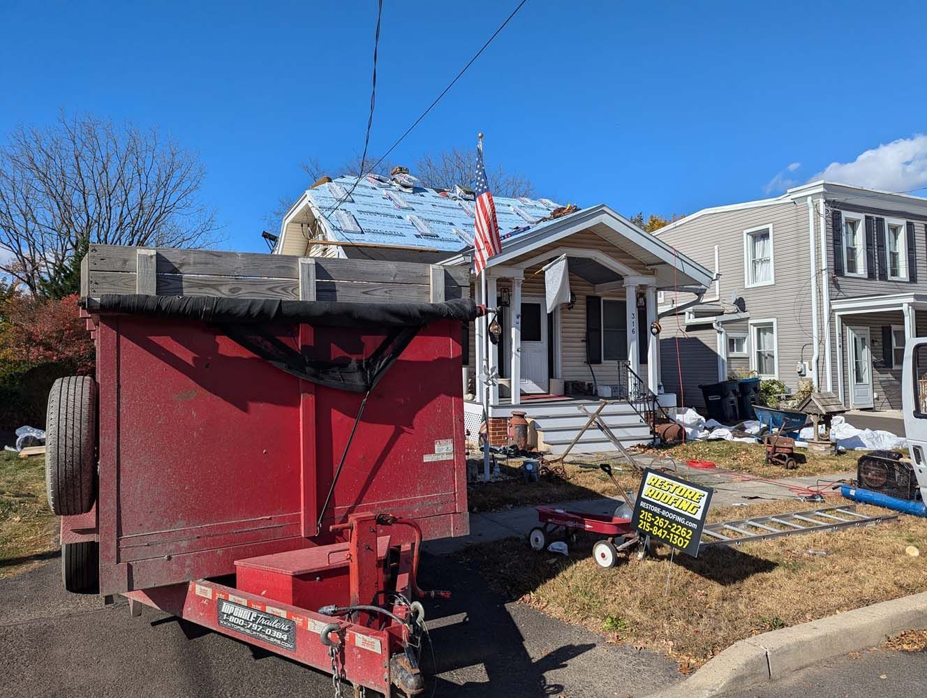 Red dumpster trailer in front of a house with roofing in progress on a sunny day.
