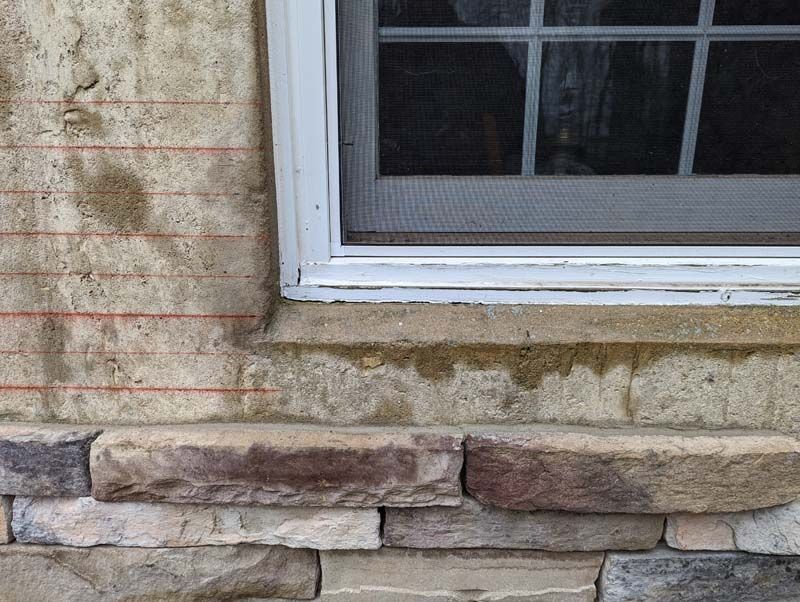 Close-up of a building exterior with stone veneer and a window, showing water stains and exposed building materials.