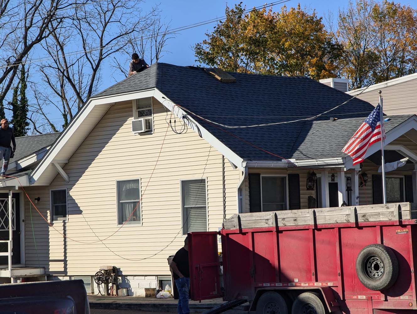 Workers on a house roof replacing shingles; red trailer in foreground, American flag on the porch.