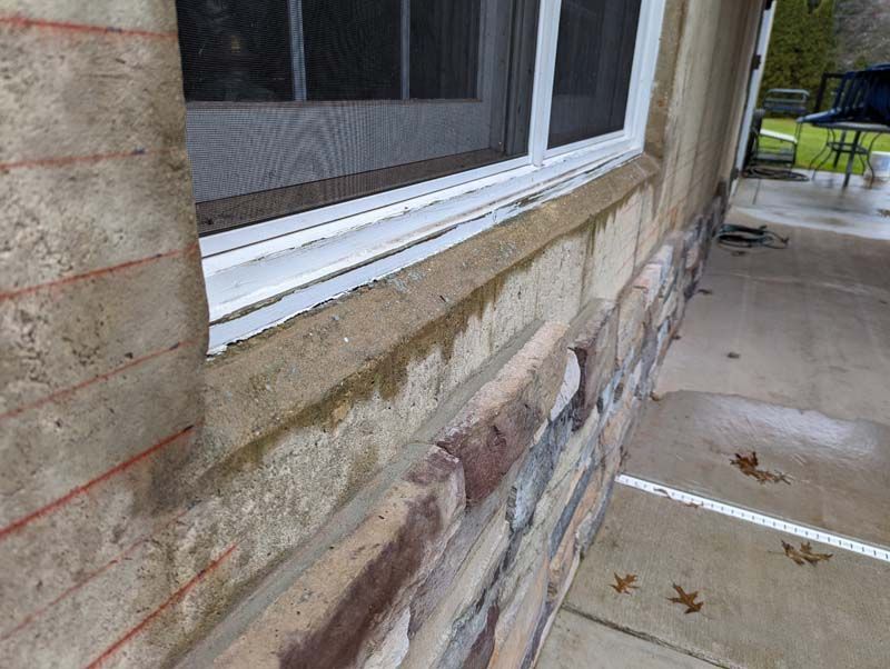 Exterior view of a window with stone and stucco wall, with dampness visible.