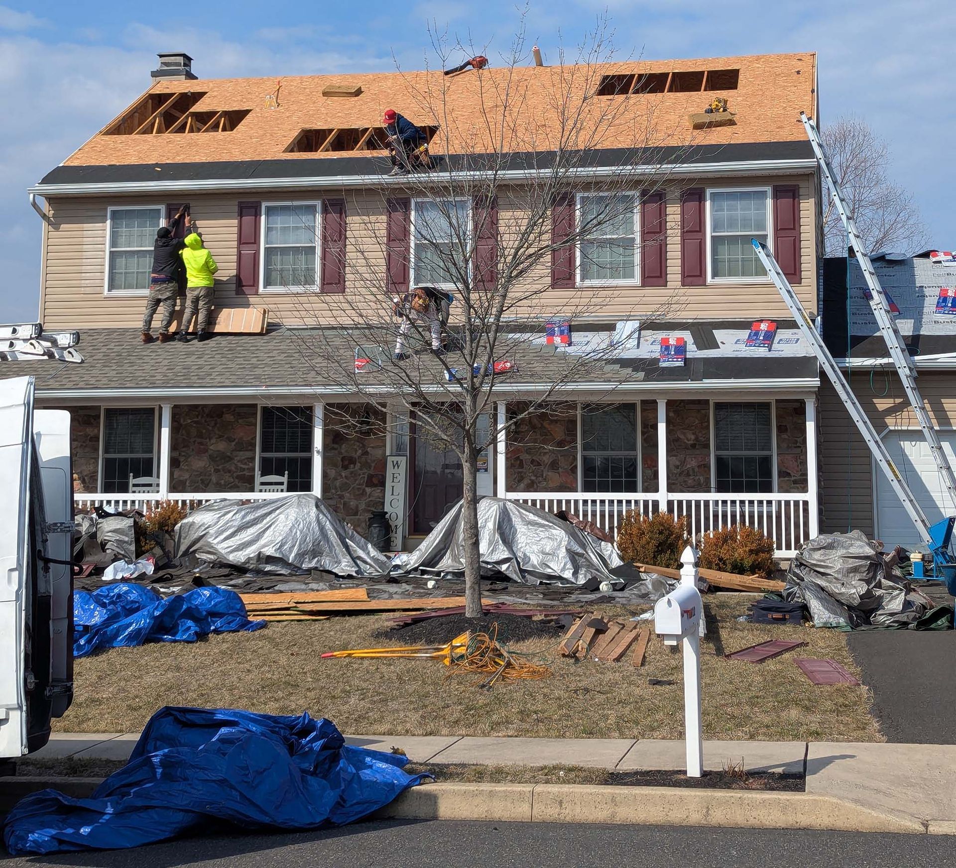 Workers replacing a roof on a two-story house with brown siding. Tarps cover the yard, and a ladder is propped against the roof.