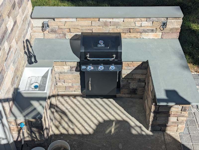 Outdoor kitchen with grill, sink, and stone facade. Grey countertop. Shadowed patio.