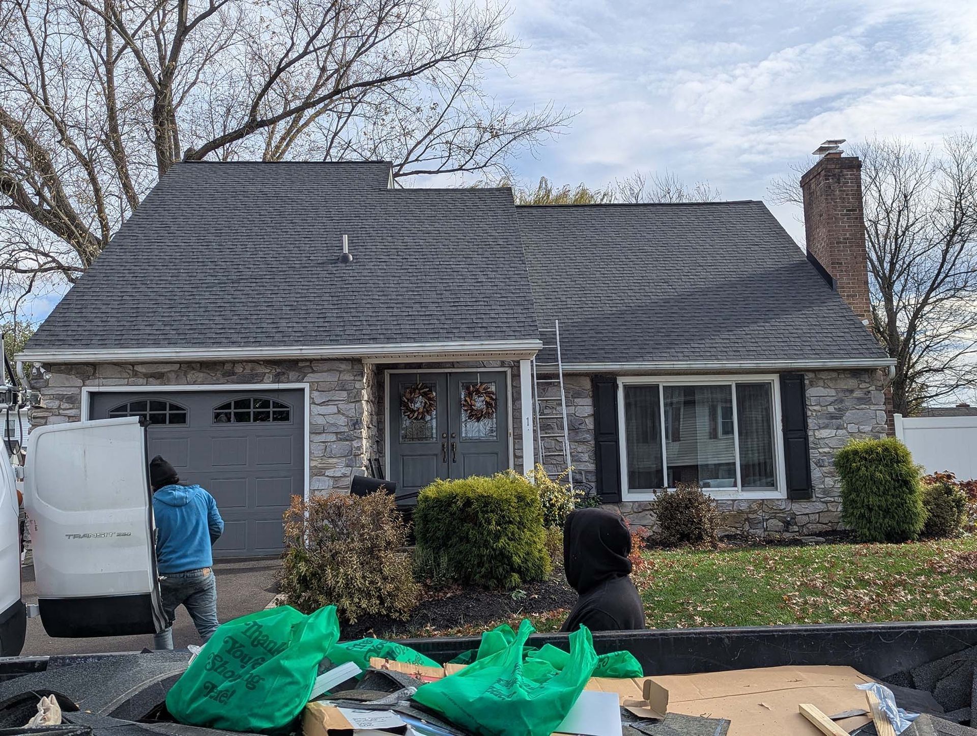 Workers unloading debris from a dumpster near a gray-shingled house with stone facade, garage, and chimney.